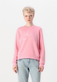 Pink sweatshirt made from soft material, featuring a crew neck, embroidered logo, and relaxed fit, paired with light wash jeans.