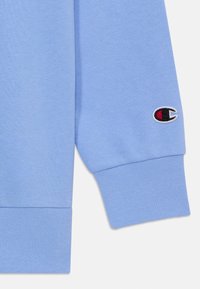 Tissu de sweatshirt bleu clair avec des poignets et un ourlet côtelés. Présente un logo brodé avec des détails rouges et noirs sur la manche gauche.