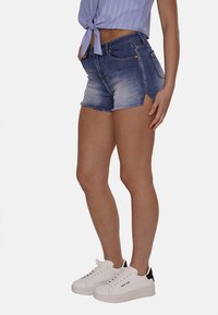 Shorts in denim con orlo sfilacciato, gradient da blu chiaro a blu scuro, con tasche frontali e un design aderente, abbinati a sneakers bianche.