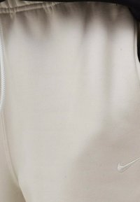 Ljusbeige Nike-joggingbyxor med elastisk midja, textur som liknar mjuk bomull, med en liten vit logotyp på nedre högra sidan.