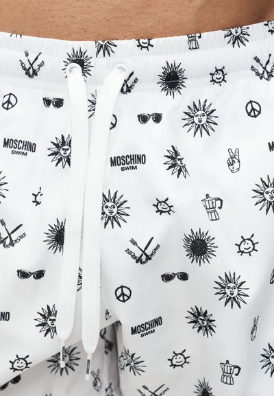 Hvite badeshorts med svarte solansikter, fredstegn, solbriller, kryssede gafler og kniver, kaffetraktere og "Moschino Swim"-logoer, med hvite snorer.