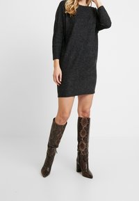 Robe pull oversize grise à manches longues, associée �à des bottes hautes en cuir marron foncé avec un motif serpent et des talons compensés.