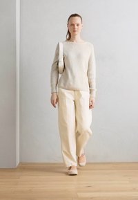 Maglione a maglia beige chiaro, pantaloni a gamba larga color crema e scarpe slip-on rosa tenue. La modella porta una borsa rotonda di colore chiaro. Ambientazione naturale.