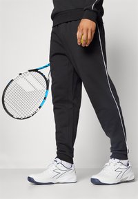 Sorte joggebukser med hvite side-striper, hvite athletic sko og et tennisracket. Joggebuksene er laget av et mykt stoff; skoene har teksturerte såler.