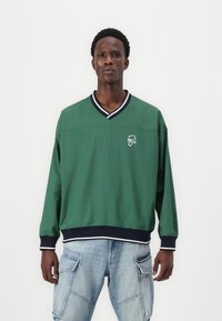 Pulover verde cu guler în V, bordură bleumarin și un logo pe pieptul stâng. Fabricat din material ușor, are mâneci lungi și o croială confortabilă.