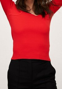 Rode geribde langodil met een diepe V-halslijn en een aansluitende silhouet, gecombineerd met zwarte broek.