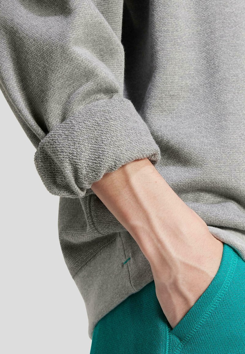 Grå sweatshirt med rullad ärm, texturerad tyg, och gröna byxor. Synlig hand i fickan, som framhäver plaggets passform och material.