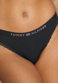 Černé tanga s krajkovým lemem, vybavené širokým elastickým pasem s nápisem "TOMMY HILFIGER" v bílém písmu. Měkká, hladká látka.