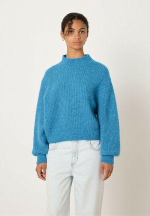 Suéter turquesa con cuello alto, textura acanalada y hombros caídos. Lleva puesto sobre unos jeans azul claro, un poco holgados. Tejido suave y de punto.