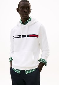 Man wearing a white Tommy Hilfiger hoodie over a green and white striped shirt with navy pants, hands in pockets, looking ahead.