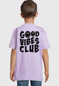 Lila īsām piedurknēm T-krekls ar lieliem melniem burtiem, kas lasāmi "GOOD VIBES CLUB", un diviem smaidošiem sejiņām uz aizmugures, izgatavots no mīkstas auduma.