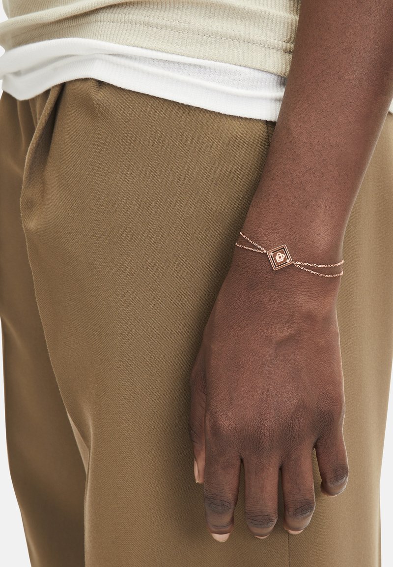 Rose gold bracelet with a square pendant, featuring delicate chain links. Worn on a hand resting against brown trousers.