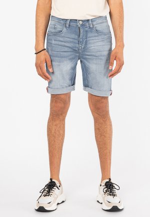 Helle blaue Jeansshorts mit klassischem Schnitt, umgeschlagenen Säumen, Vordertaschen und dezentem Ausbleichen. Getragen mit weißen und beigen Sneakers.