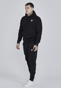 Black hoodie with front pocket and white logo, paired with matching joggers. Features ribbed cuffs, soft texture, worn with white sneakers.
