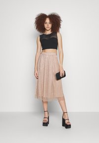 Musta lyhyt crop-top, jossa on läpikuultava paneeli, yhdistettynä polvipituiseen vaaleanpunaiseen kimaltavaan hameeseen. Malli pitää mustaa clutch-laukkua ja käyttää mustia korkokenkiä.