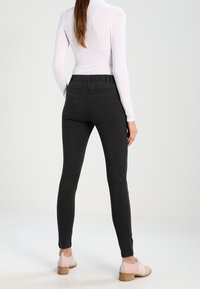 Donkergrijze leggings met een taps toelopende pasvorm, elastische tailleband en gestructureerde stof, gecombineerd met lichtroze schoenen op een gladde ondergrond.