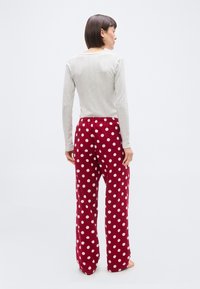 Γυναίκα που στέκεται ξυπόλητη με την πλάτη γυρισμένη, φοράει ανοιχτόχρωμο μακρυμάνικο μπλουζάκι και κόκκινο παντελόνι πιτζάμα με λευκές πουά.