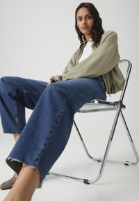 Woman with long dark hair wearing an olive jacket, white shirt, and wide-leg blue jeans, sitting on a clear folding chair against white background.