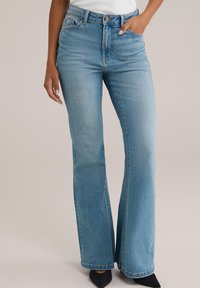 Hellblaue Schlagjeans aus Denim, mit hoher Taille, Fünf-Taschen-Design und leichter Ausbleichung, kombiniert mit schwarzen Spitzenschuhen.