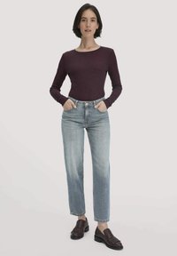 Frau steht mit den Händen in den Taschen, trägt ein enges langärmliges dunkelviolettes Oberteil, helle blaue Jeans und dunkelbraune Mokassins.