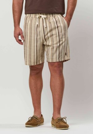 Man die beige en bruin gestreepte korte broek met trekkoord draagt, een bruin shirt en beige veterschoenen, staand met handen in de zakken.