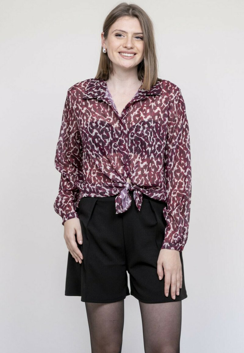 Blouse bordeaux transparente avec un imprimé léopard, dotée d'une taille nouée et d'une fermeture à boutons, associée à un short noir taille haute.