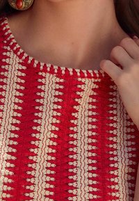 Textil bordado en rojo y crema con un patrón geométrico. Cuello redondo con costuras decorativas. Modelo tocando el hombro.