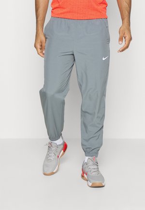 Homme portant un pantalon de jogging Nike gris, des baskets grises et blanches avec des accents rouges, et un t-shirt rouge, debout devant un fond uni.