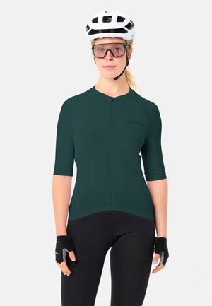 Femme portant un casque de vélo blanc, des lunettes de cyclisme teintées, un maillot à manches courtes vert foncé, des gants de cyclisme noirs et un pantalon de cyclisme noir.