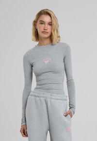 Grå langermet topp laget av mykt stoff, med en tettsittende design og en liten rosa sommerfugl-logo på brystet.