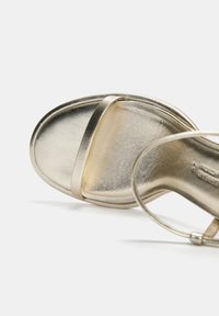 Sandalo con tacco metallizzato oro, dalla superficie liscia, con una fascia singola sopra le dita dei piedi e una cinghia alla caviglia con un piccolo dettaglio a fibbia.