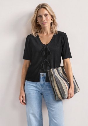 Kvinde med skulderlangt blond hår bærer en sort bluse med bindefront, lyseblå jeans, gyldne blomsterøreringe og holder en stribet mulepose.