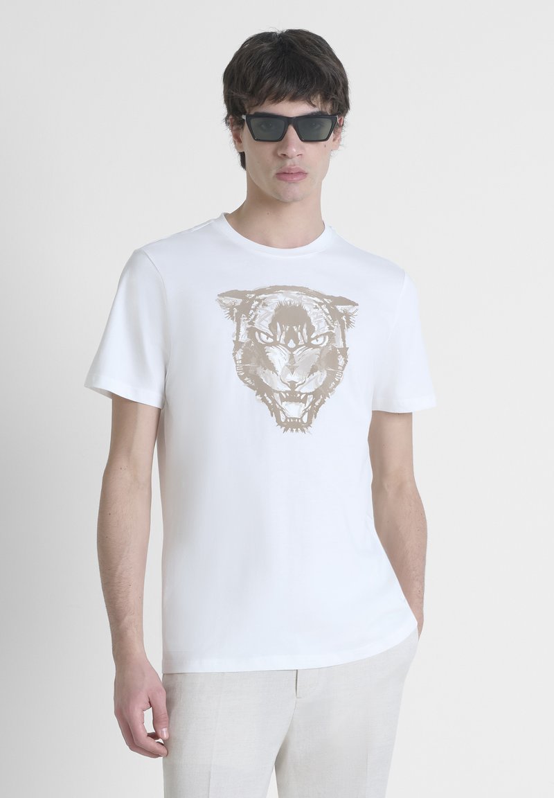 Camiseta de algodón blanca con un gran gráfico de tigre beige en la parte frontal. Manga corta y cuello redondo. El modelo lleva gafas de sol oscuras.