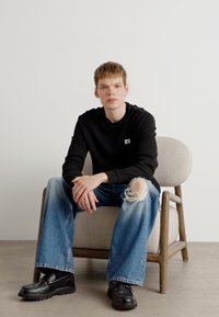 Zwarte geribbelde lange mouwen shirt, lichtblauwe versleten wijde jeans en zwarte chunky schoenen. Model zittend in een lichtgekleurde stoel.