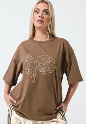 Femme aux cheveux blonds lisses portant un T-shirt marron oversize avec des contours de coquillages blancs, associé à un pantalon crème à motifs de coquillages, mains dans les poches.