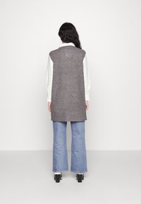 Henkilö seisoo selkä kameraan päin, päällä pitkä harmaa hihaton villapaita, valkoinen paita, vaaleansiniset farkut ja mustat korot.