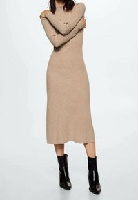 Robe beige tricotée à manches longues et silhouette cintrée, présentant une texture lisse et des détails côtelés, associée à des bottines noires.