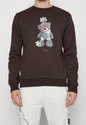 Persona que lleva una sudadera marrón con un gráfico de un oso de peluche gris, ojos rojos, una corona blanca sobre su cabeza y pantalones blancos con cremalleras.