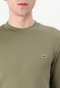 Sudadera de algodón en verde oliva con cuello redondo, que presenta un pequeño logo de cocodrilo bordado en el lado izquierdo del pecho. Textura suave.