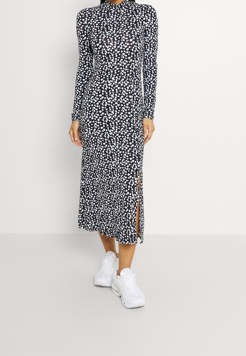 Femme portant une robe midi à manches longues à pois noirs et blancs avec une fente latérale, associée à des baskets blanches épaisses.