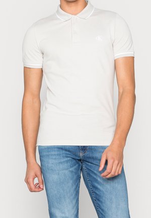 Polo crème avec bordure blanche, manches courtes et petit logo CK. Porté avec un jean bleu délavé. Texture de tissu lisse.