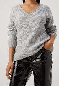Maglione grigio chiaro lavorato a maglia con scollo a V e maniche lunghe arrotolate; abbinato a pantaloni in finta pelle nera ad alta lucentezza.