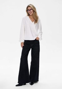 Weißes Langarm-V-Ausschnitt-Oberteil mit gerafften Bündchen, kombiniert mit weit geschnittenen schwarzen Jeans und schwarzen Absatzstiefeln. Auf einem schlichten hellen Hintergrund.