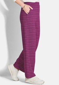 Persoon in paarse met patronen broek, witte sneakers, witte sokken, een witte top en een gouden kettingarmband met de hand in de broekzak.