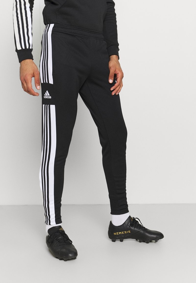 Person trägt schwarze Adidas-Trainingshose mit weißen Streifen und schwarze Stollenschuhe mit der Aufschrift "Nemesis", steht vor einem schlichten hellen Hintergrund.