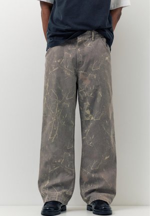 Osoba nosiaca voľné nohavice s maskáčovým vzorom v kombinácii s čiernymi topánkami a zastrčenou námorníckomodrou košeľou, stojaca pred jednoduchým sivým pozadím.