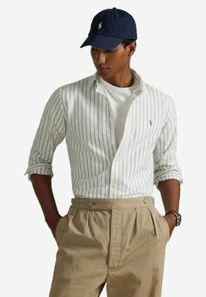 Chemise à rayures blanches et bleues, portée ouverte sur un t-shirt blanc, associée à un pantalon plissé beige et une casquette de baseball marine.
