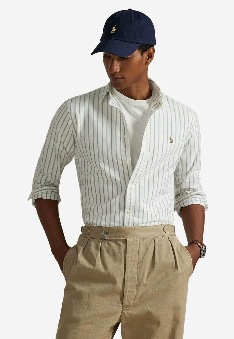 Chemise à rayures blanches et bleues, portée ouverte sur un t-shirt blanc, associée à un pantalon plissé beige et une casquette de baseball marine.