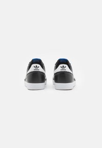 Svarta lädersneakers med vita gummisulor, blå detaljer på hälen och ett distinkt tre-stripes logotypdetalj på baksidan.