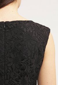 Vestido negro de encaje con un patrón floral, diseño sin mangas, cuello alto y cierre de cremallera en la parte de atrás. Se aprecian detalles de tela texturizada.